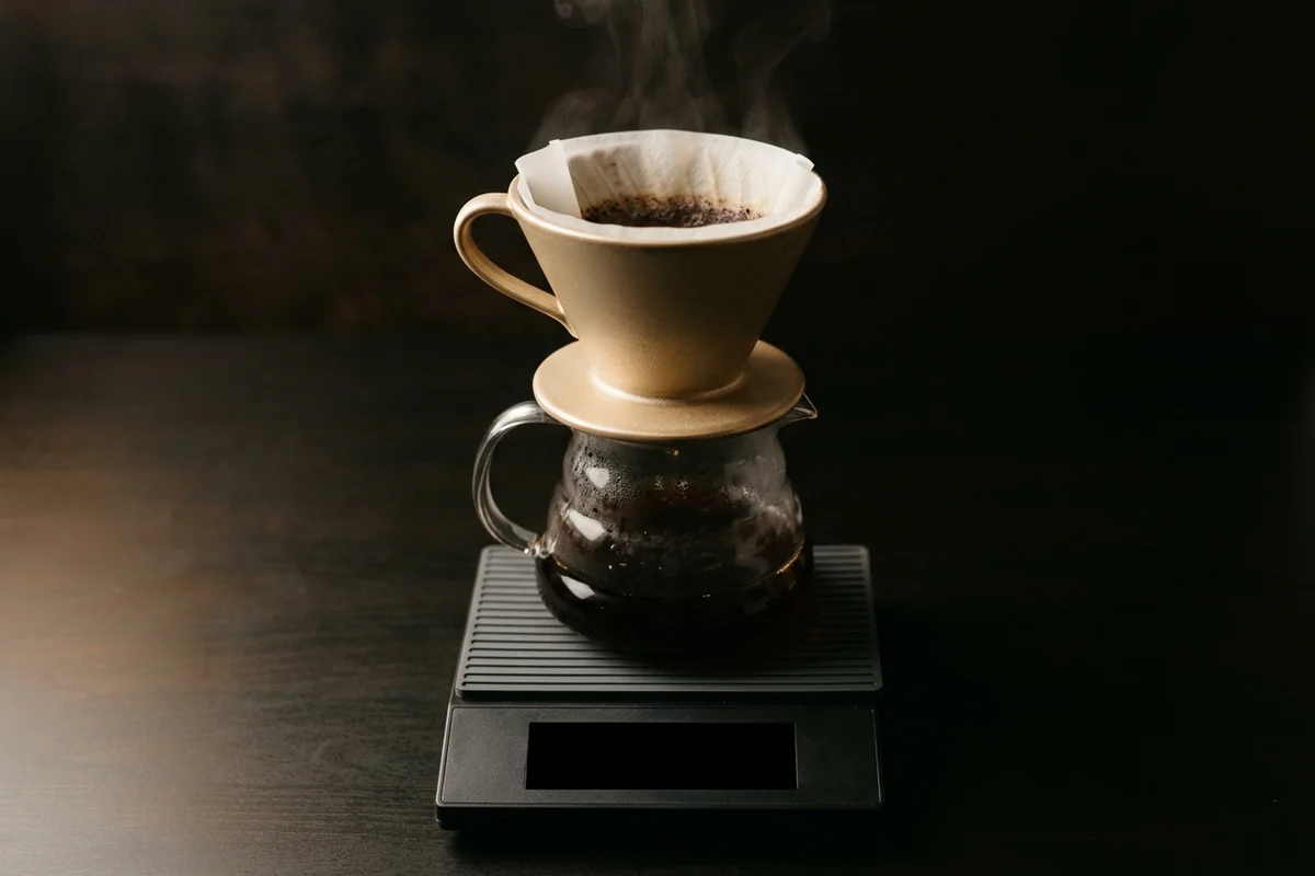 A ceramic pour-over cone and glass server standing on a flat slab scale with a thin silicone pad between them, steam rising from the cone, the scale's display a blank dark panel.