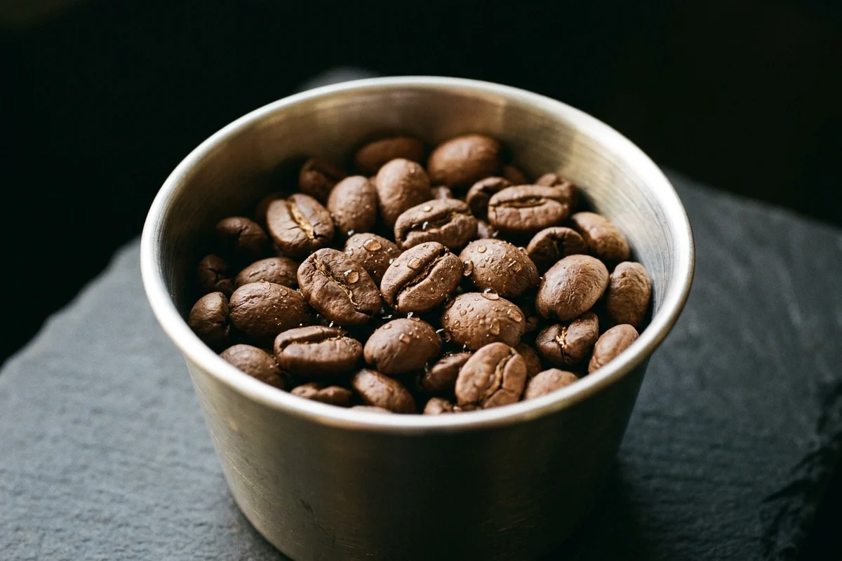 Whole roasted coffee beans in a plain metal dosing cup, fine water droplets clinging to their surfaces and caught in the key light, the stone under the cup completely dry.