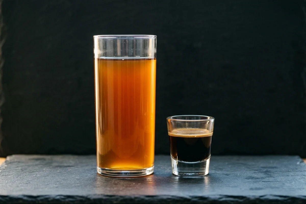A tall glass of translucent amber filter coffee standing beside a small thick-walled espresso glass holding a near-opaque dark shot with a crema collar.
