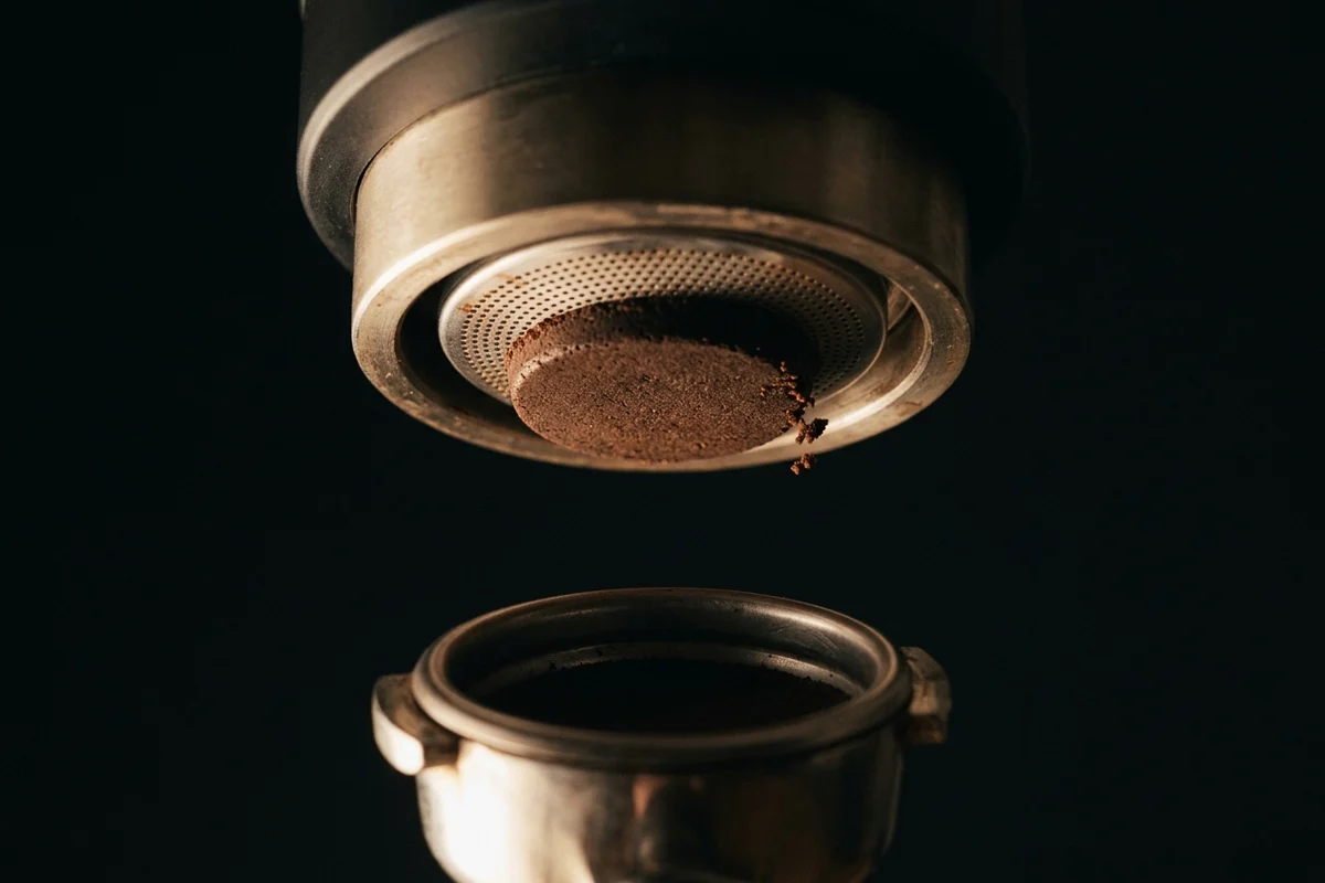 The underside of an espresso group head seen from below with a spent coffee puck stuck flat against the perforated shower screen, a few crumbs dropping from one edge, and the empty portafilter basket below and out of focus.