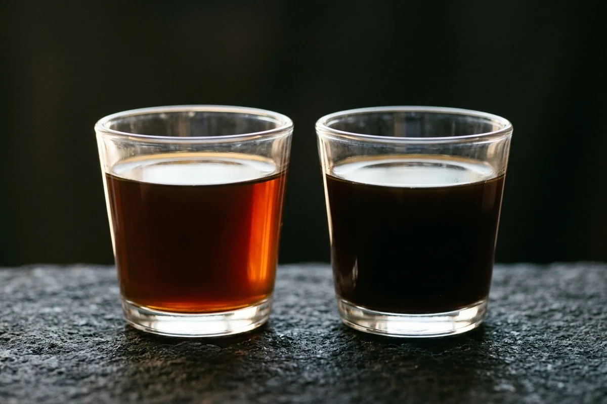 Two identical tumblers side by side on dark stone, filled to exactly the same level, the left holding a translucent amber-red brew lit from behind and the right a nearly opaque dark one.